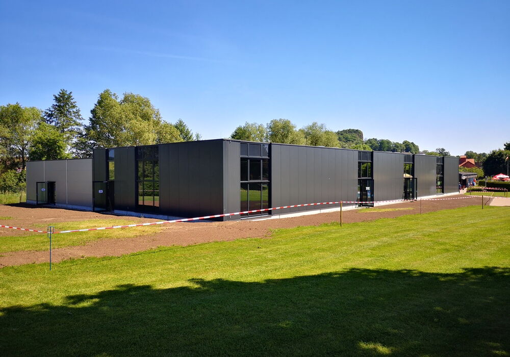 Ein modernes, graues Gebäude mit großen Fensterflächen steht auf einer Wiese unter blauem Himmel